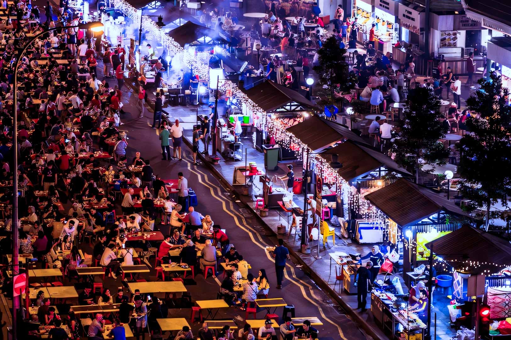 Aerial night view of a vibrant outdoor food street filled with dining tables, food stalls decorated with string lights, and crowds enjoying local street food.