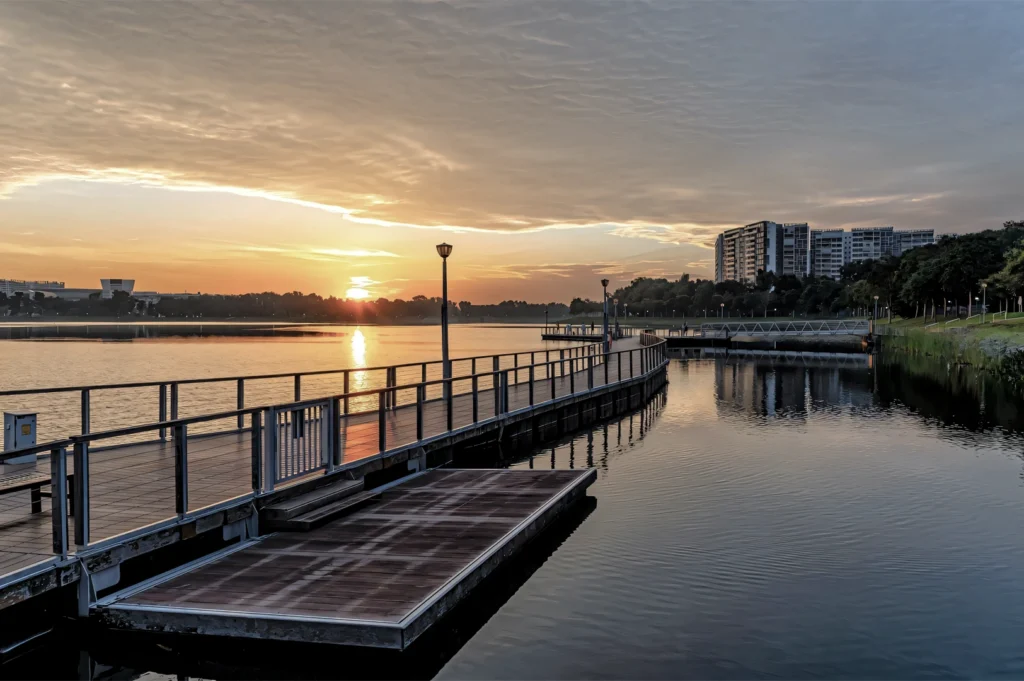 Scenic sunset over Bedok Reservoir with a wooden pier, still water reflections, and residential buildings in the distance.