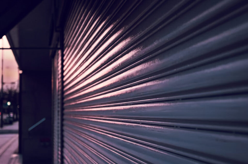 Closed metallic storefront shutter with gradient lighting at dusk, highlighting linear pattern and urban street backdrop.