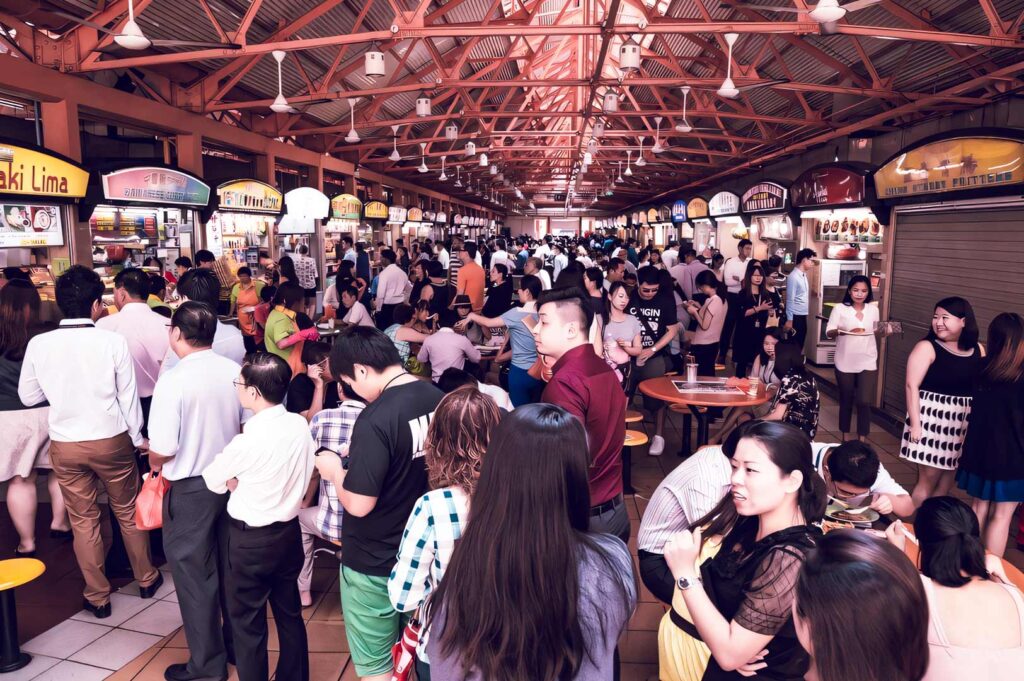 A bustling hawker centre filled with people lining up at various food stalls under a high metal roof with bright signage and busy dining tables.