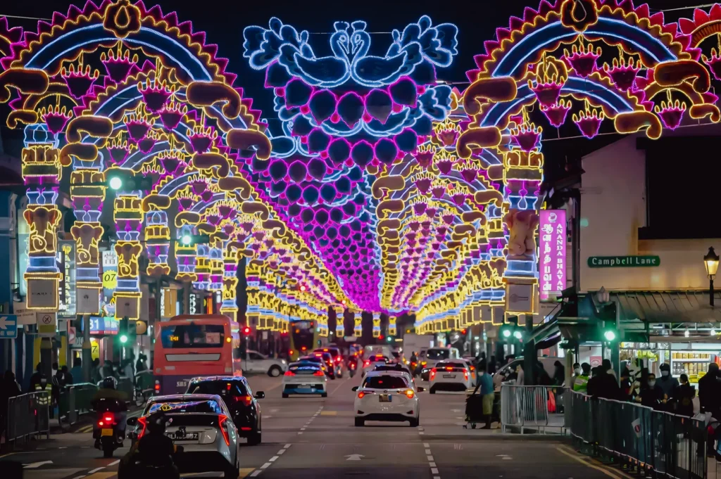 Wide-angle eye-level night shot of a city street lined with colorful illuminated archway decorations, moving traffic, and pedestrians along barricades, showcasing festive street lights and urban celebration atmosphere.