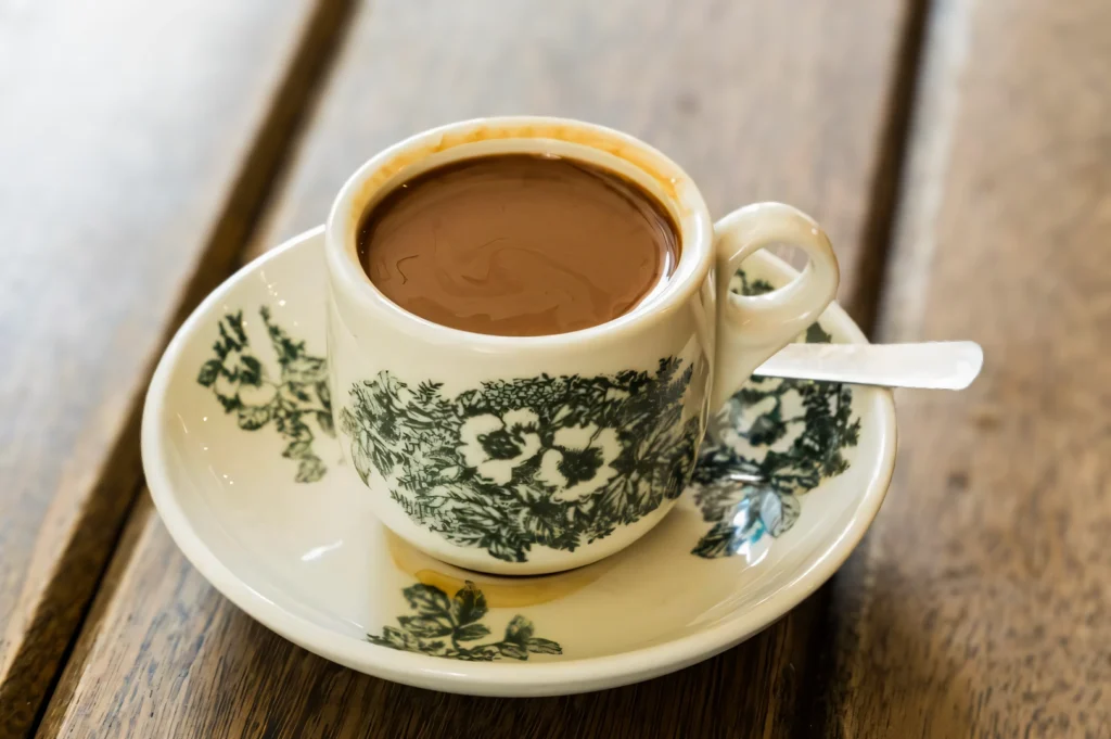 Close‑up of a classic kopitiam coffee served in a vintage green‑floral ceramic cup and saucer with a teaspoon on a wooden table.