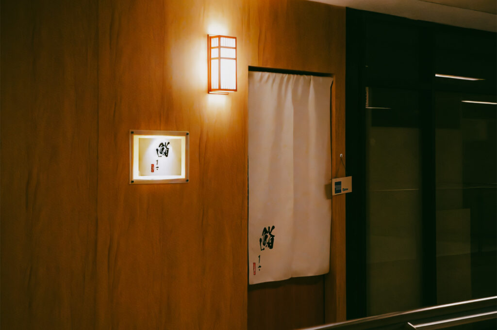A Japanese restaurant entrance with a wooden façade, soft wall lighting, and a white noren curtain featuring traditional calligraphy.