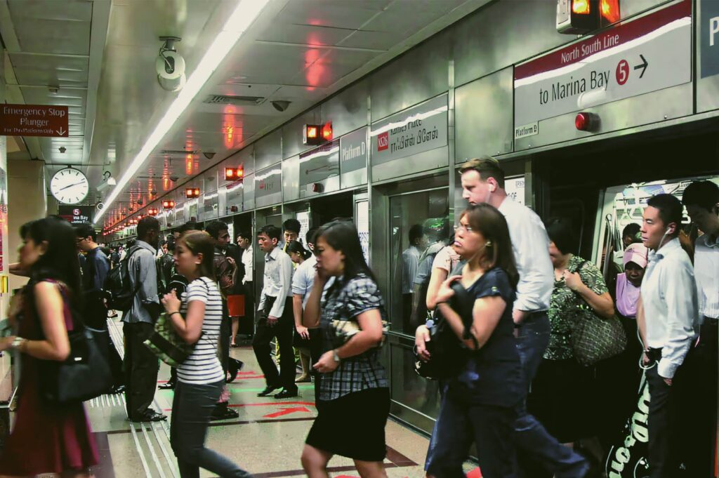 A busy MRT platform with commuters entering and exiting the train on the North South Line, with station signs and platform doors visible.