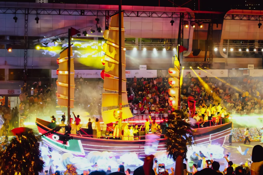 Wide-angle elevated shot of a brightly illuminated festival parade featuring a large traditional-style boat float with tall sails, stage lighting, smoke effects, and a packed indoor arena filled with spectators celebrating a cultural event.