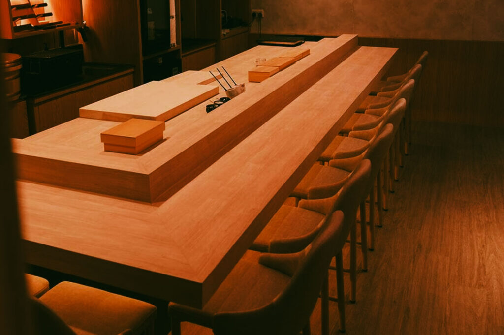 A minimalist Japanese sushi counter with warm wooden interiors, soft ambient lighting, and neatly arranged cushioned chairs surrounding a central chef’s workstation.
