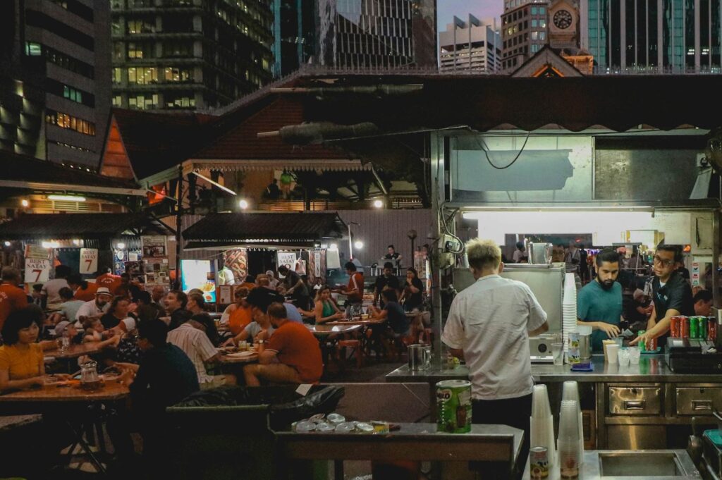 An outdoor hawker centre surrounded by city skyscrapers, with diners eating at packed tables and a food stall preparing drinks in the foreground.