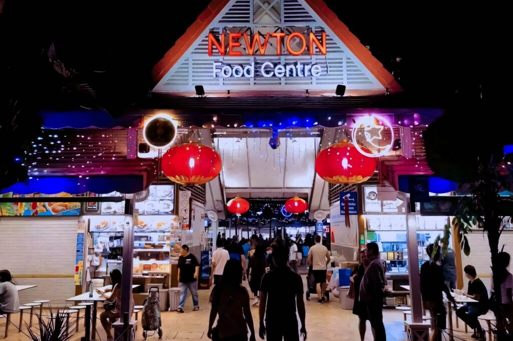 Night view of Newton Food Centre entrance illuminated with colorful lights and red lanterns, with crowds walking toward the busy hawker stalls inside.