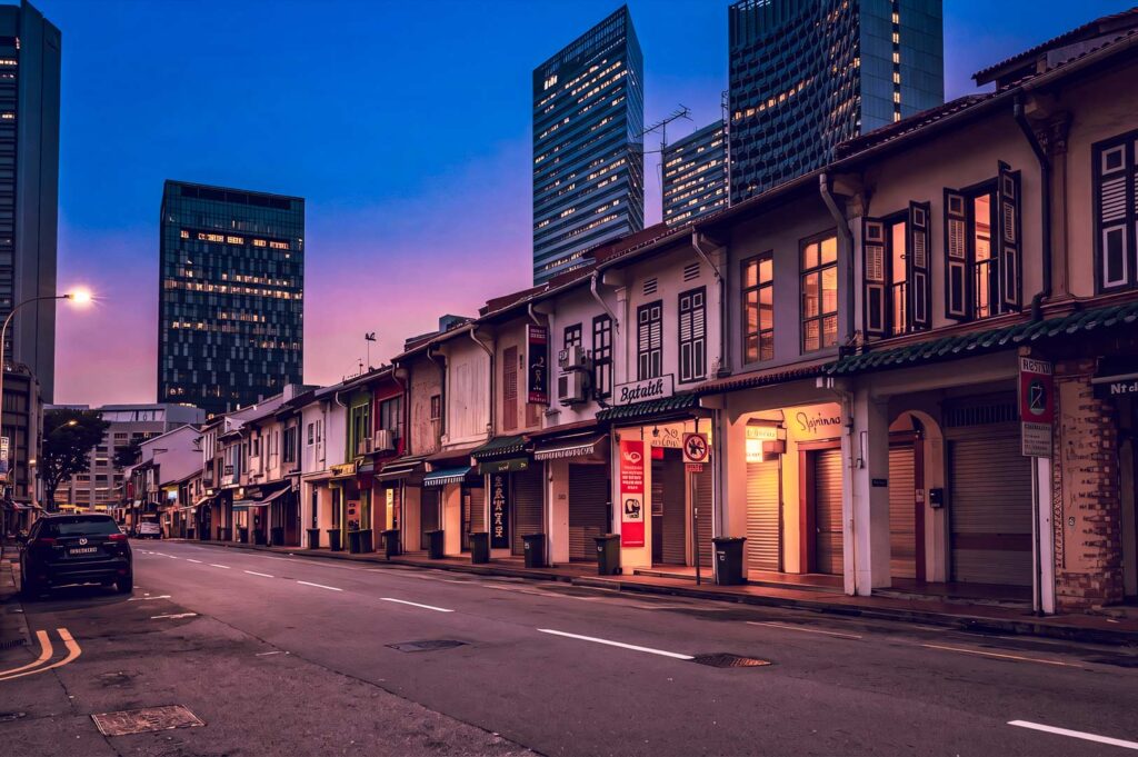 Dusk city street lined with shuttered colonial shophouses, parked car, streetlights, and skyscrapers under a pink‑blue sky.