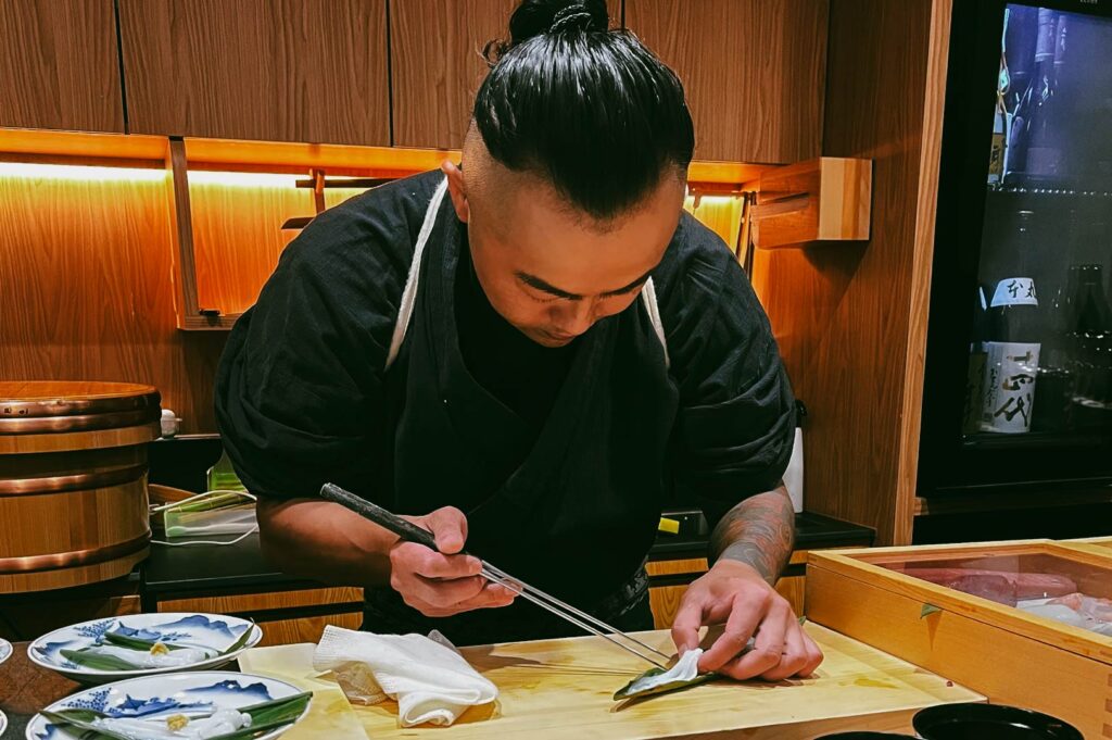 A sushi chef in traditional attire meticulously slicing fresh fish on a wooden counter inside an intimate Japanese omakase restaurant.”