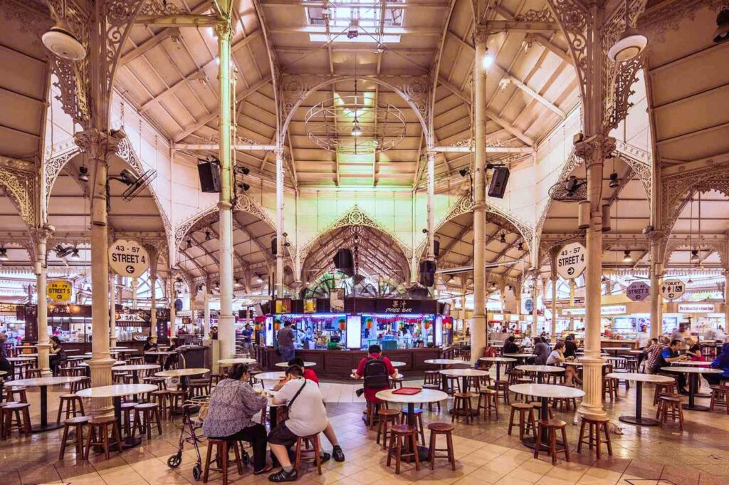 Wide interior view of Lau Pa Sat with ornate cast‑iron columns, high arched ceilings, and rows of tables surrounding the central food stalls.