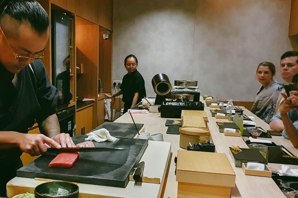 A Japanese omakase dining setup with a chef cutting premium tuna behind a wooden counter as guests observe the preparation of handcrafted sushi.