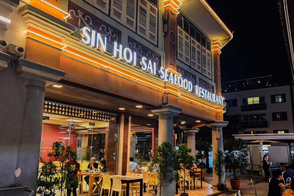 Night-time view of Sin Hoi Sai Seafood Restaurant with warm exterior lighting, outdoor seating, and diners visible inside the brightly illuminated eatery.