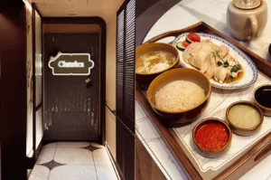 Left: Wide-angle eye-level entrance shot of Chatterbox restaurant in Singapore, featuring a modern corridor design with warm lighting, decorative wall panels, patterned floor tiles, and a glowing Chatterbox sign creating an upscale dining atmosphere; Right: Top-down close-up shot of a premium Singapore Hainanese chicken rice set served on a wooden tray, showcasing sliced poached chicken with light soy sauce, fragrant rice, clear chicken soup, chili sauce, ginger sauce, and condiments arranged neatly in ceramic bowls.