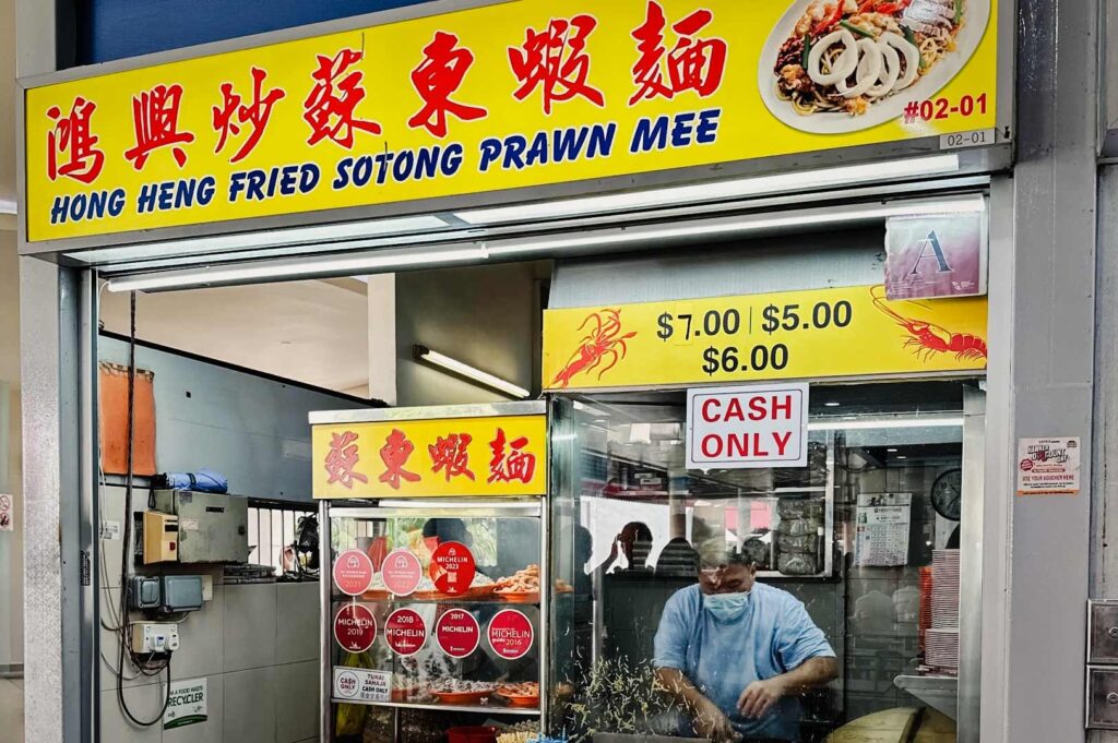 Front view of the Hong Heng Fried Sotong Prawn Mee stall at Tiong Bahru Market, featuring a bright yellow signboard, menu prices, and a cook preparing food inside the hawker stall.