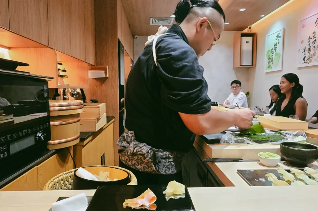 Close-up, eye-level shot of a sushi chef preparing fresh sashimi and ingredients at a Japanese omakase counter in a modern minimalist restaurant, highlighting authentic culinary craftsmanship and premium dining experience.
