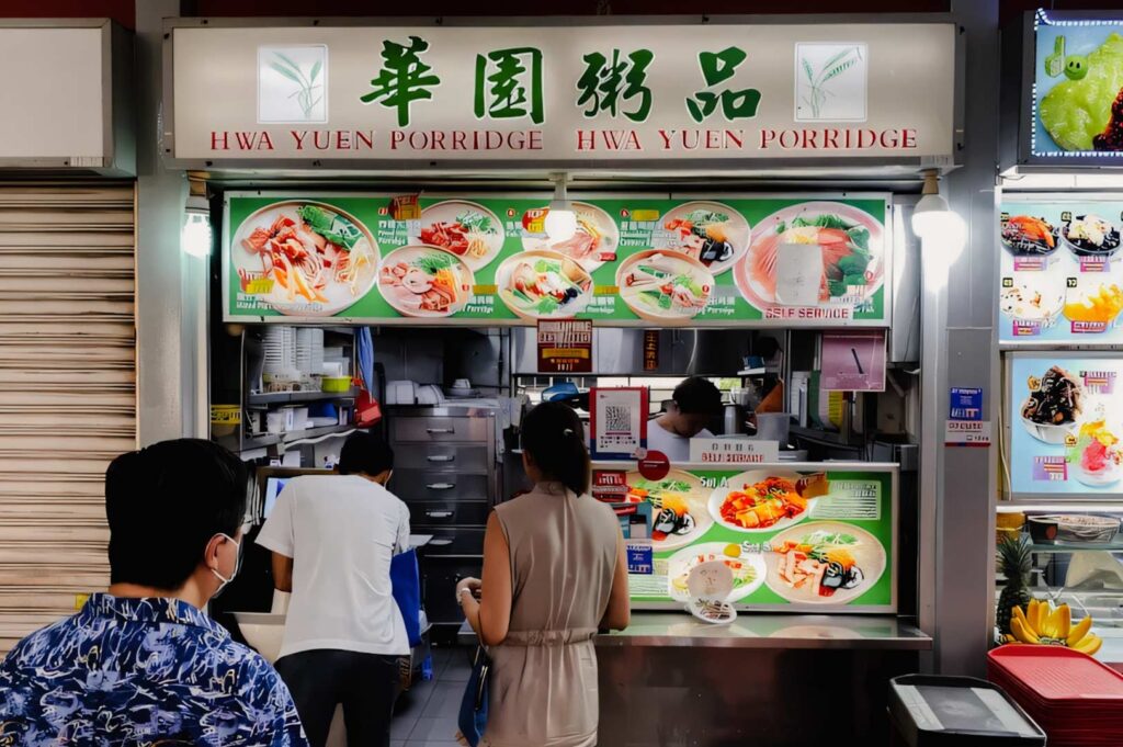 Hwa Yuen Porridge stall at Tiong Bahru Market displaying colorful menu boards with various porridge dishes, with customers queuing at the counter.