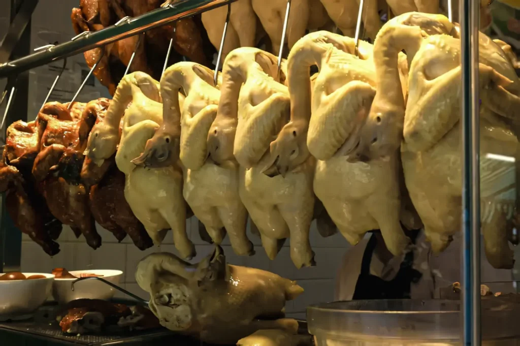 Eye‑level market stall shot showing rows of boiled and roasted ducks hanging on hooks, with trays of braised eggs beneath, evoking the traditional preparation methods that accompany dishes like Hainanese chicken rice in Chinese and Southeast Asian cuisine.