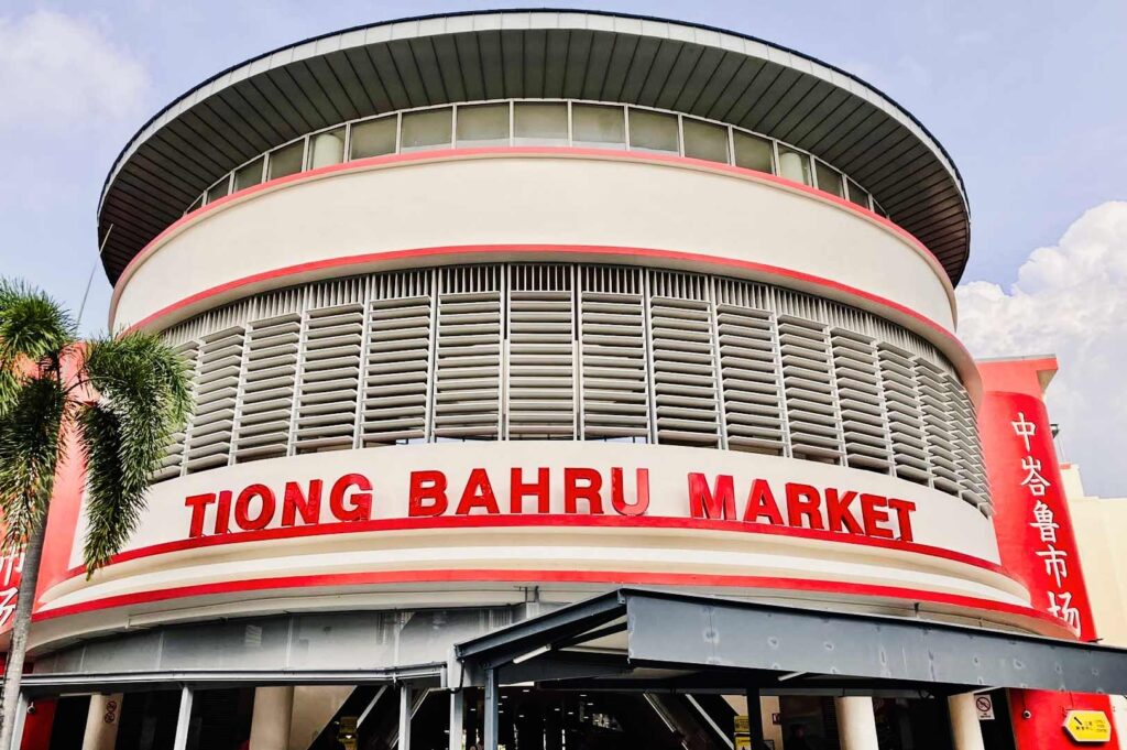 Exterior view of Tiong Bahru Market featuring its curved modern façade, large red signage, and surrounding palm trees under a clear sky.
