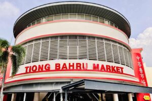Exterior view of Tiong Bahru Market featuring its curved modern façade, large red signage, and surrounding palm trees under a clear sky.