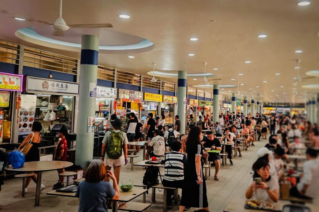 Busy interior of Tiong Bahru Market hawker centre with numerous food stalls, round communal tables, and diners enjoying meals in an open, brightly lit space.