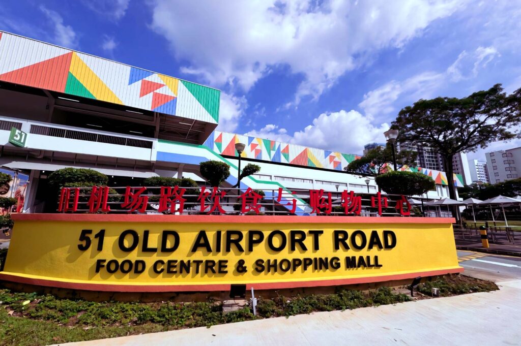 Front facade of 51 Old Airport Road Food Centre & Shopping Mall, featuring a bright yellow sign, geometric building patterns, and a clear blue sky.