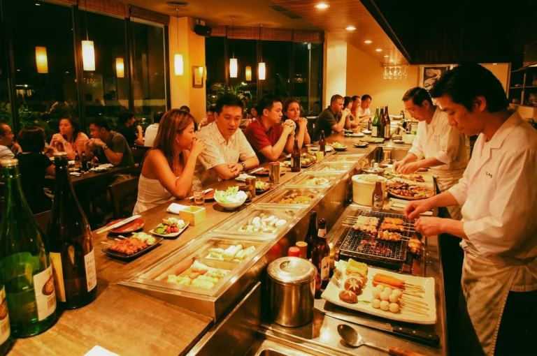 Wide eye-level angle of a lively izakaya restaurant with diners seated around an open kitchen counter while chefs grill skewers, highlighting authentic Japanese dining atmosphere