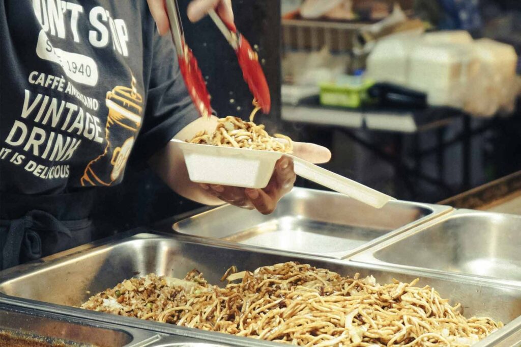 Food vendor using tongs to pack freshly cooked noodles into a disposable takeaway container at a food stall.