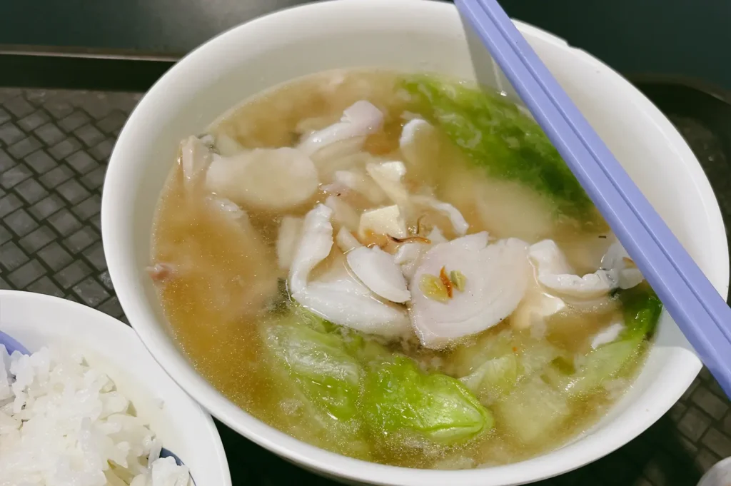 Close-up, top-down angle shot of a bowl of clear broth soup with sliced fish and leafy vegetables, served with chopsticks on top and a side bowl of white rice.
