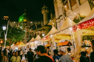 Wide-angle night street shot of a bustling Ramadan bazaar in Kampong Glam, Singapore, with festive lights, food stalls, and crowds enjoying seasonal festival food.