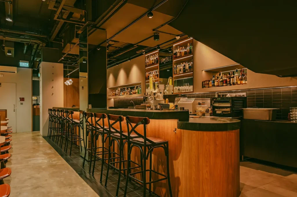 Wide-angle perspective of a modern Singapore bar with wooden counters, high stools, warm lighting, and a sleek contemporary interior.