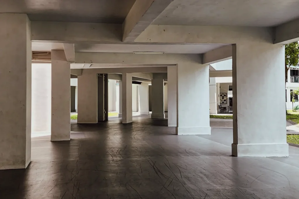 Low‑angle wide shot of an open concrete void deck with structural columns, clean lines, and natural light, showcasing minimalist urban architecture and flexible community spaces commonly found in Singapore neighborhoods.