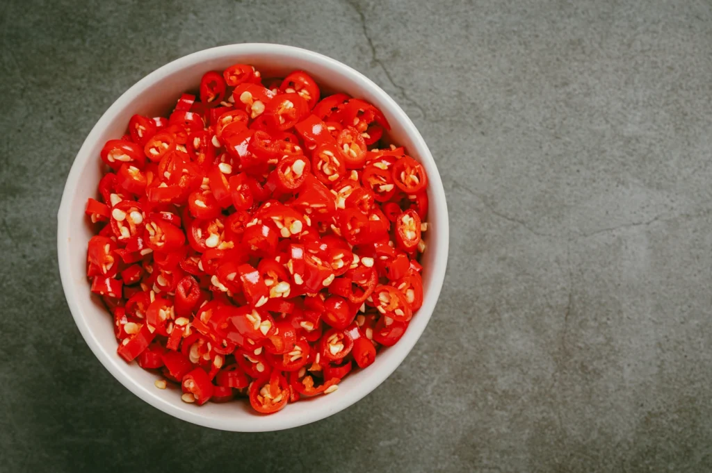 Overhead top-down shot of a white bowl filled with sliced red chili peppers, showing seeds and vibrant texture on a gray stone background.