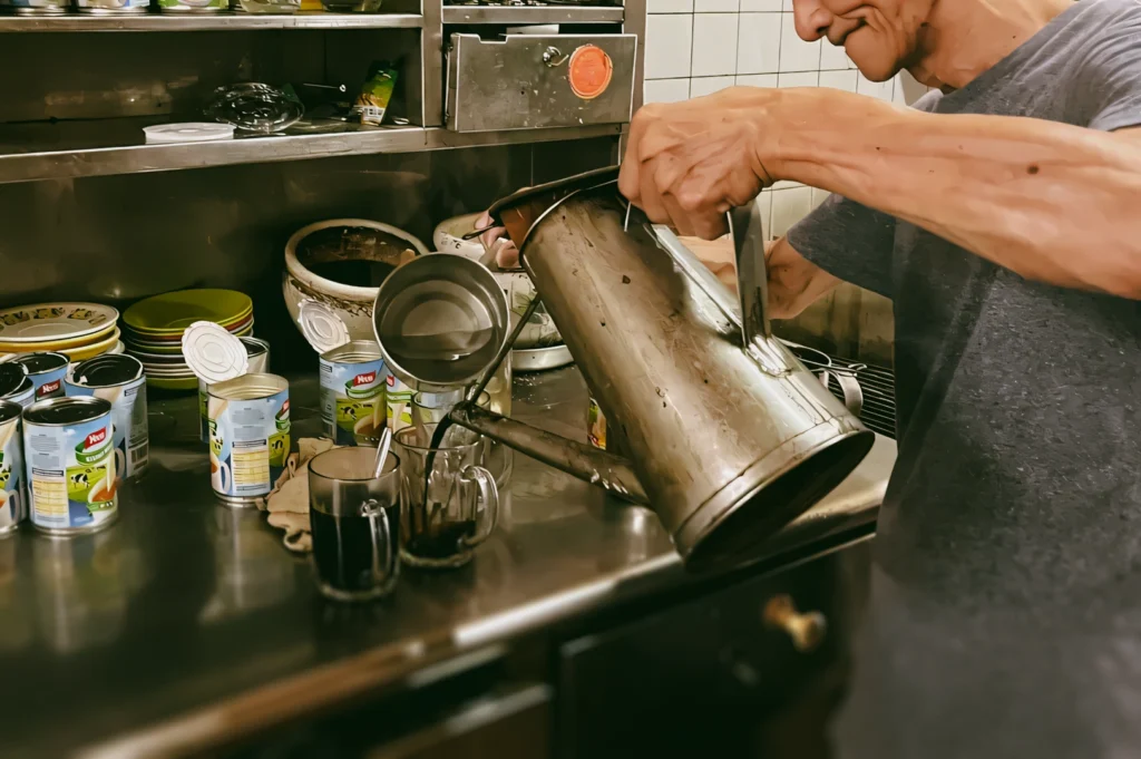 Close side-angle shot of hands pouring coffee from a vintage metal kettle into glass mugs on a café-style stainless steel countertop.