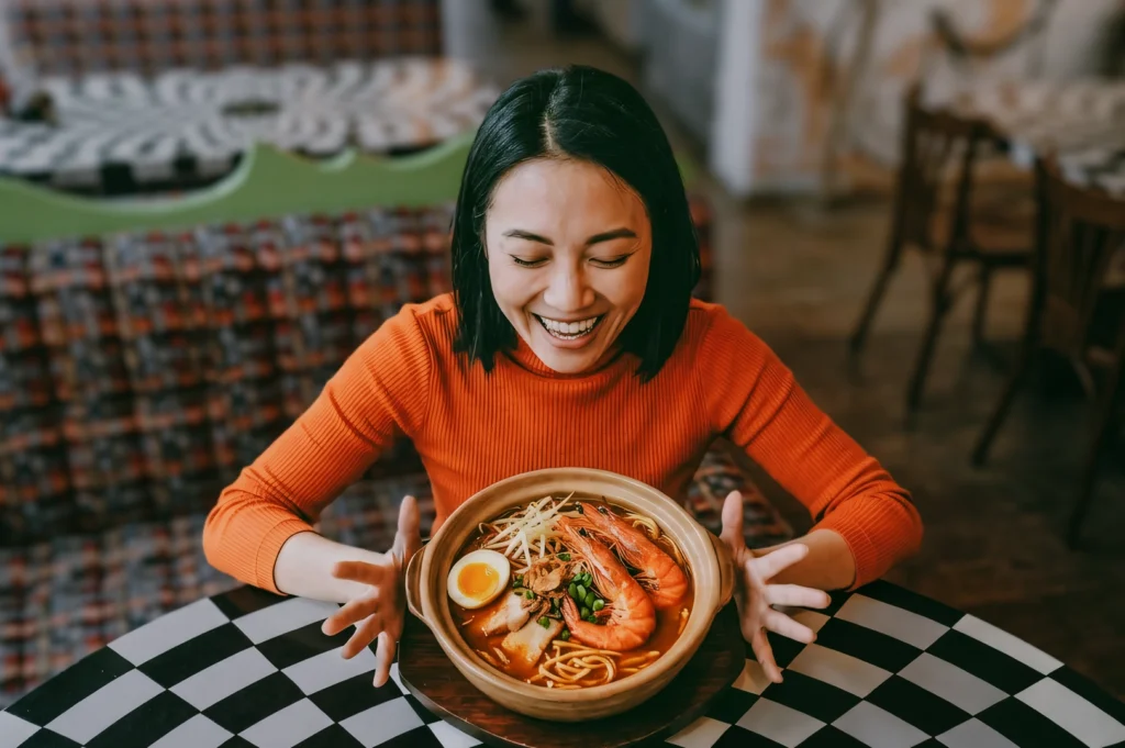 Close-up front high-angle shot of a person seated at a restaurant table holding a bowl of prawn noodle soup with shrimp, egg, and noodles, showcasing Singapore hawker food culture and the joy of traveling for a favorite meal