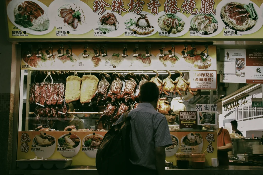 Front-facing wide shot of a Chinese roast meat stall displaying hanging char siu, roast duck, and roast pork beneath a menu board with Chinese text, as a customer orders at a traditional hawker center.