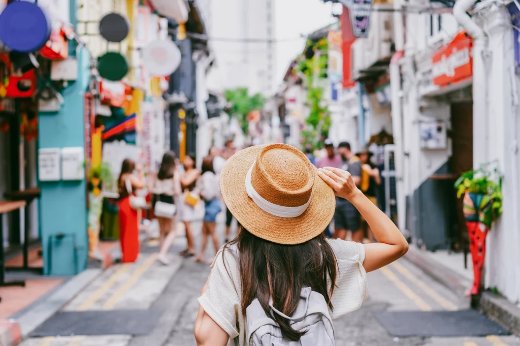 Eye-level medium-wide shot from behind of a person wearing a straw hat and backpack walking through a narrow neighborhood street, capturing the experience of wandering without a plan and discovering local food spots.