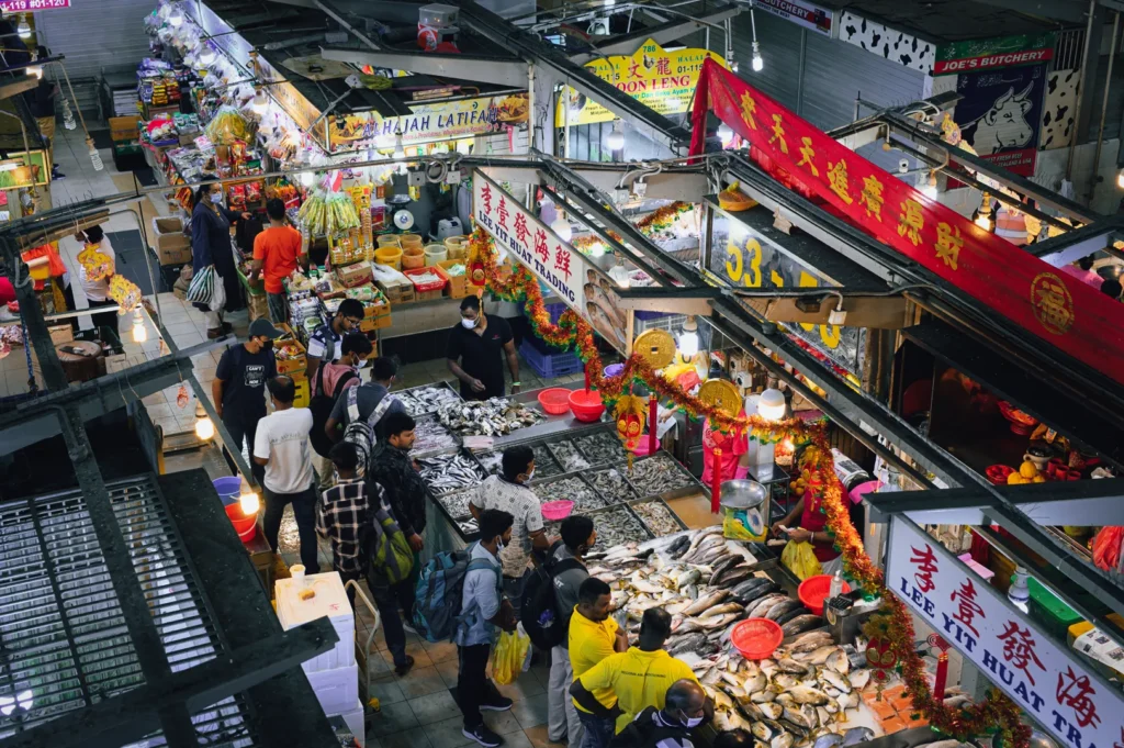 High-angle wide shot of Tekka Market in Little India, Singapore, showing bustling seafood stalls, fresh fish displays, colorful shop signs, and shoppers moving through narrow aisles.
