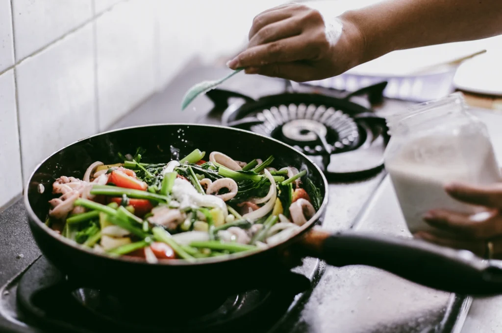 Overhead close-up shot of hands preparing a home-cooked meal by seasoning fresh vegetables in a pan on a gas stove, symbolizing comfort food, family tradition, and soul-nourishing home cooking.