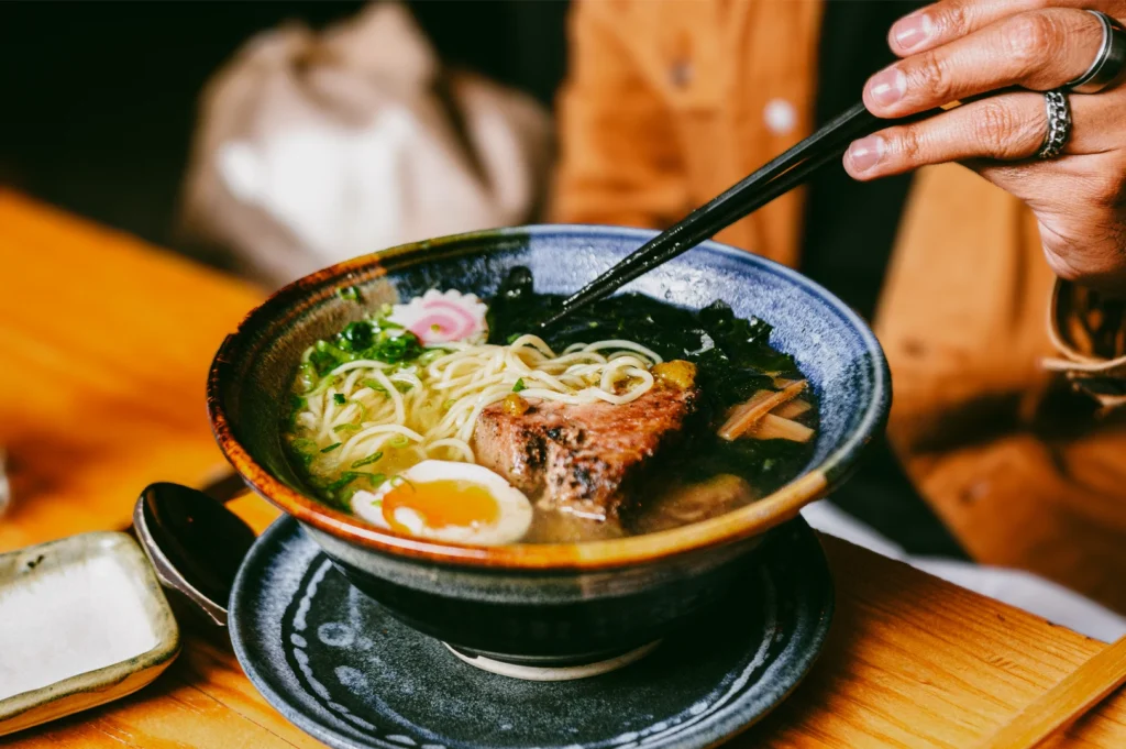 Close-up, three-quarter angle shot of a steaming bowl of Japanese ramen with noodles, soft-boiled egg, braised pork, and seaweed, as chopsticks lift noodles during a cozy restaurant dining experience.