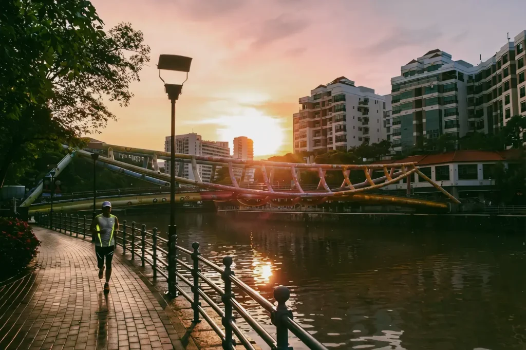 Eye-level sunrise view along the Singapore River promenade, showing a riverside walkway with railings, a pedestrian in motion, calm water reflections, and residential buildings lit by early morning light.