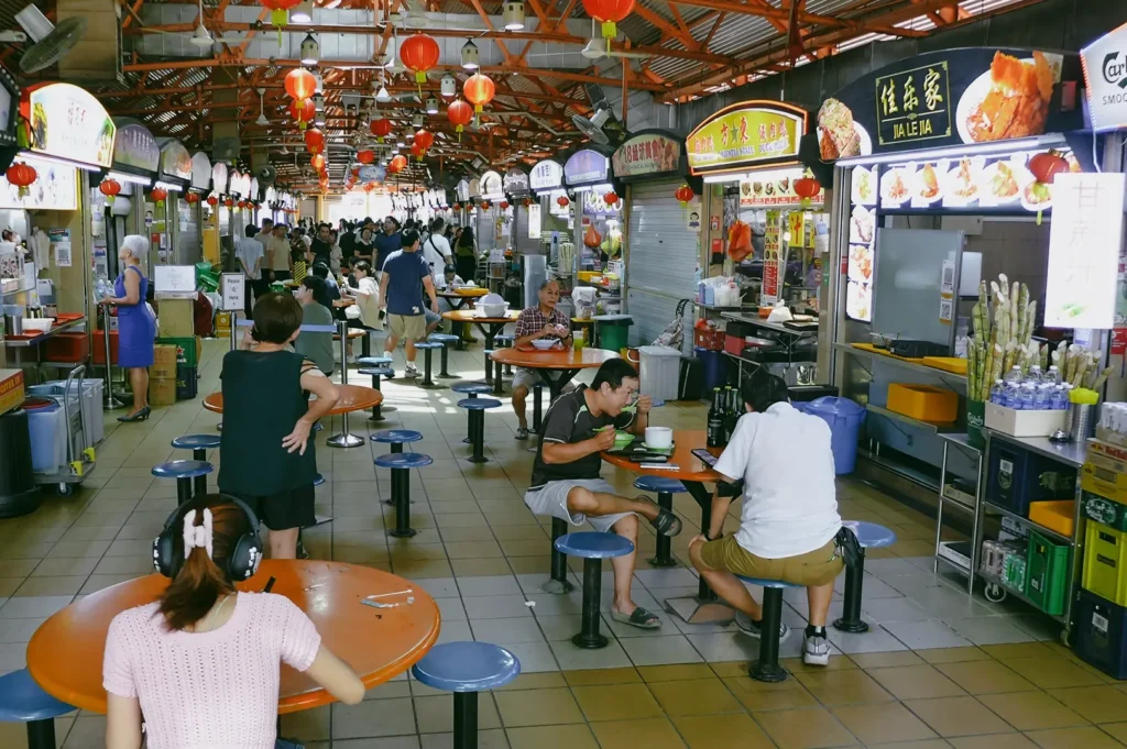 Wide‑angle eye‑level shot of a busy hawker centre with diners seated at shared round tables, surrounded by illuminated food stalls with menu signage inside a covered market hall.
