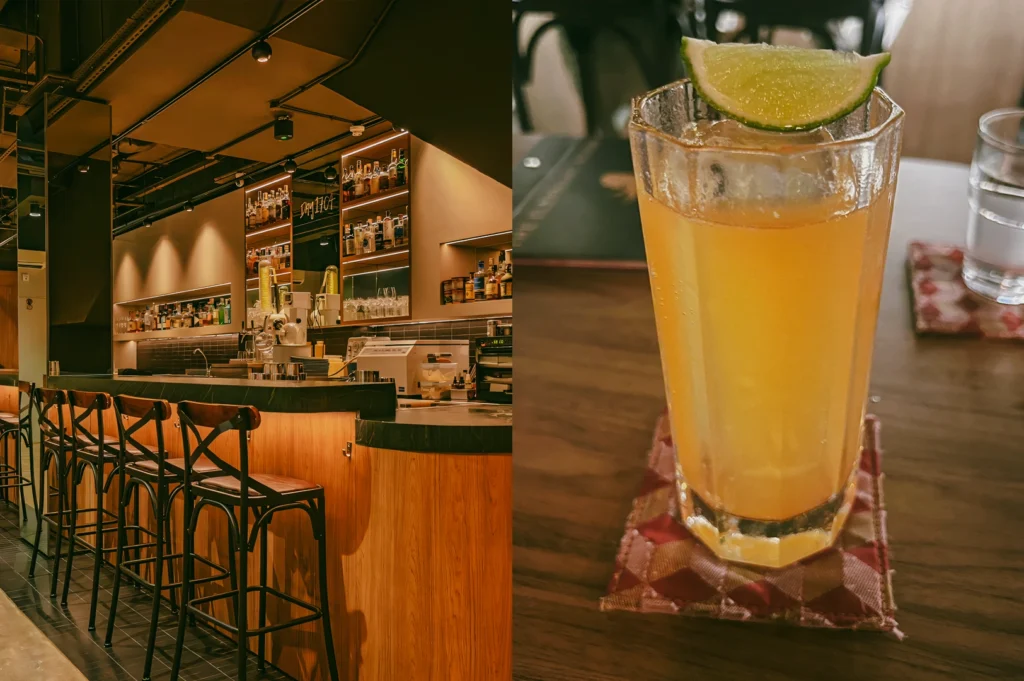 Wide-angle, eye-level view of a modern bar interior with a wooden counter, high stools, open shelving filled with liquor bottles, and warm ambient lighting highlighting textured walls and ceiling fixtures, creating a cozy, contemporary atmosphere. Right: Close-up, eye-level shot of a tall iced citrus drink in a faceted glass with a lime wedge garnish, placed on a wooden table with a folded cloth underneath, featuring soft background blur and warm lighting that emphasizes condensation and glass texture.