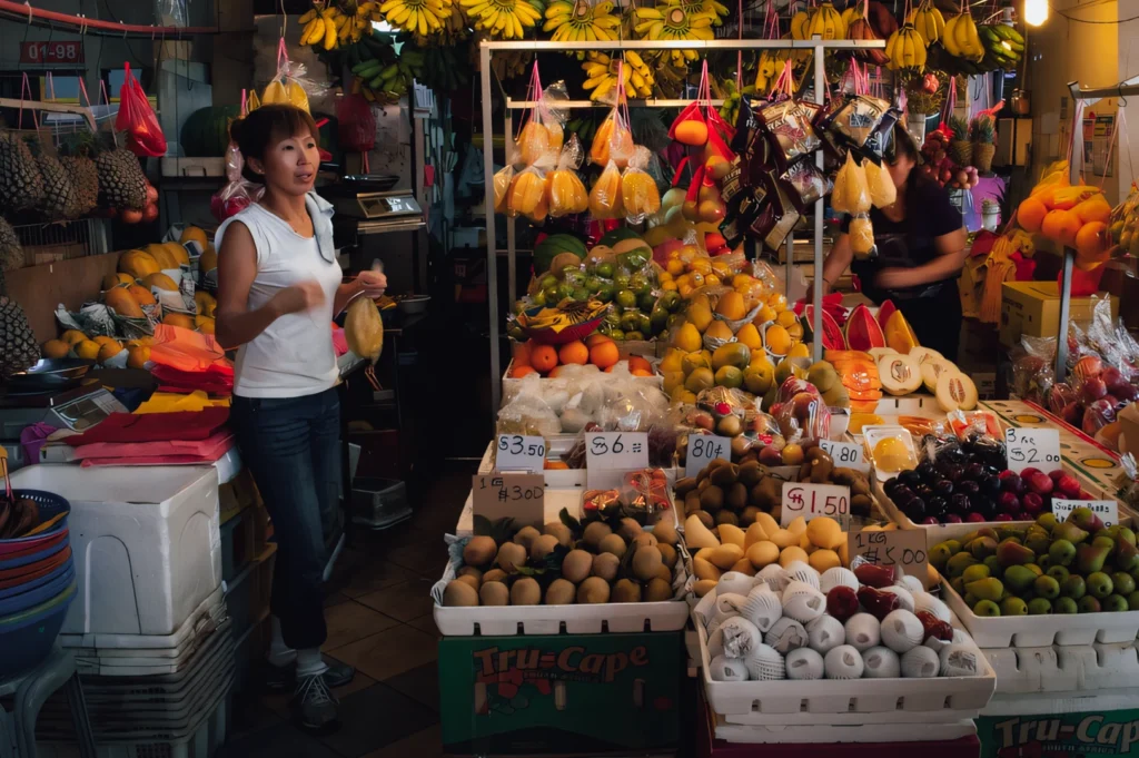 Eye-level medium-wide shot of a fruit vendor at an Asian wet market, surrounded by vibrant displays of bananas, oranges, melons, and tropical fruits with handwritten price signs.