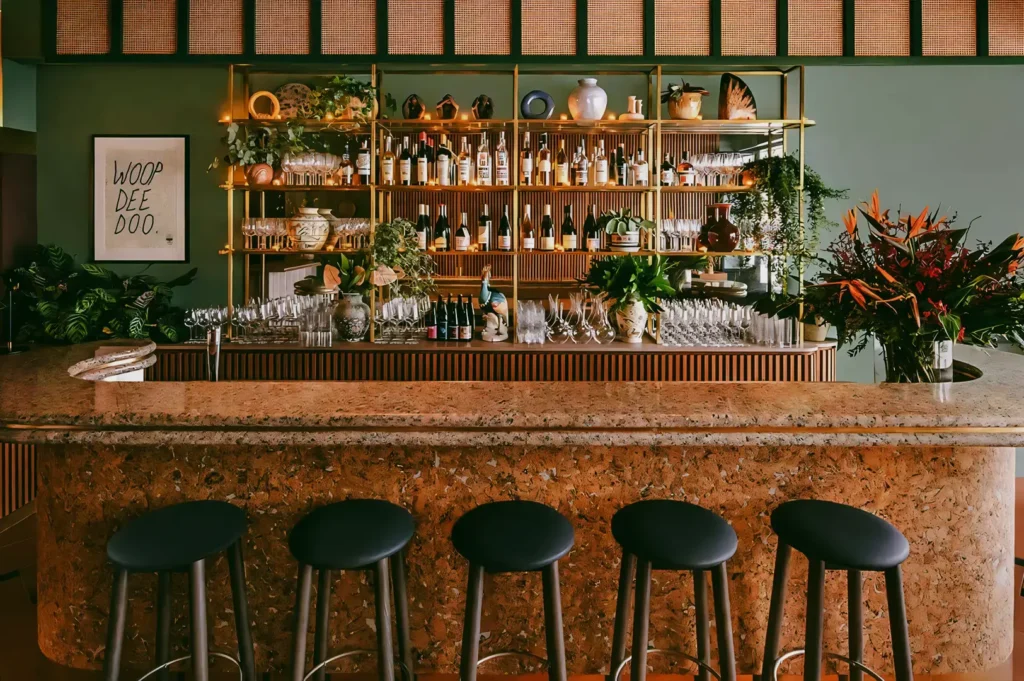 Eye-level frontal shot of a stylish modern bar in Singapore featuring a terrazzo counter, minimalist stools, lush greenery, and backlit shelves filled with bottles.