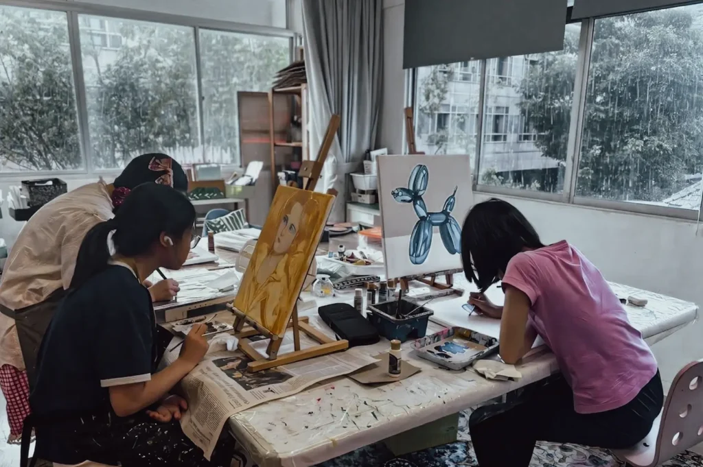 Wide‑angle interior shot of a bright art studio where participants paint on canvases at a shared table, surrounded by brushes, palettes, and artwork, showcasing creative workshops and indoor art activities.