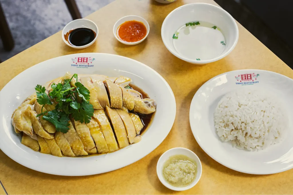 Top-down shot of a complete Kampung-style chicken rice set on a table, featuring sliced poached chicken in soy sauce, fragrant rice, clear soup, chili sauce, ginger sauce, and dark soy sauce.