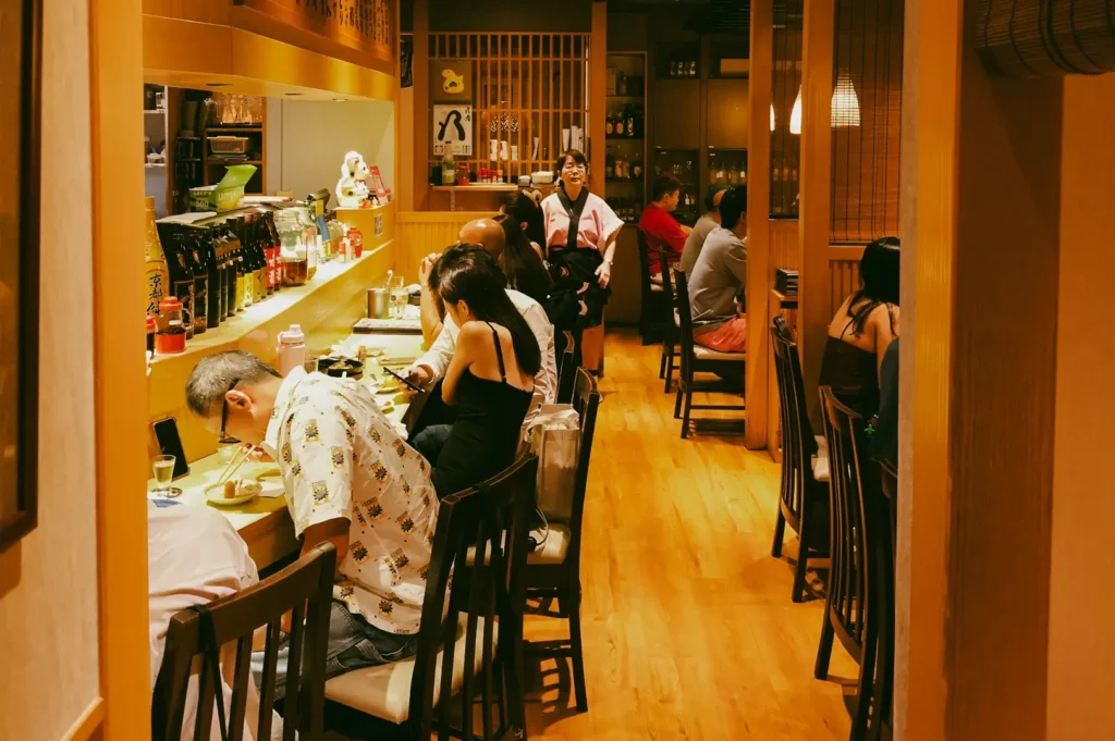 Eye‑level interior shot of a narrow Japanese restaurant at Cuppage Plaza, showing a wooden counter, bar seating, diners enjoying meals, warm lighting, and shelves stocked with bottles.