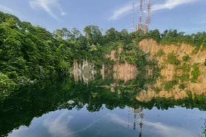 Wide-angle landscape shot of a calm quarry lake in Bukit Timah, Singapore, reflecting dense green forest, rocky cliff faces, and communication towers on the hilltop.