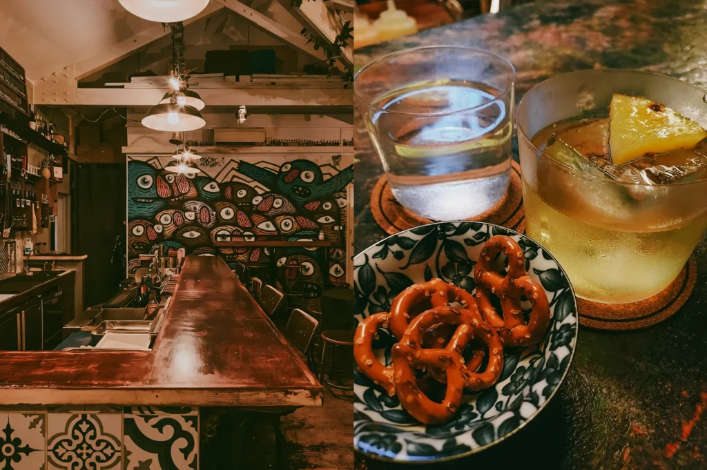 Left: An eye-level, wide-angle shot of a rustic bar interior featuring a long, polished dark wood countertop with patterned tile accents along the base. In the background, a colorful graffiti wall mural depicts abstract faces with large eyes, illuminated by warm, hanging pendant lamps suspended from an open-truss ceiling. Right: A close-up, three-quarter view of a bar setup on a textured, dark table. In the foreground, a blue and white patterned ceramic bowl is filled with salted pretzels, positioned next to a glass of iced cocktail garnished with a fresh pineapple wedge, and a clear glass of water, both sitting on cork coasters under warm, atmospheric lighting.